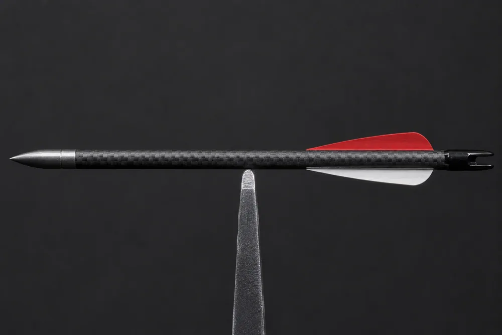 A finished carbon arrow with point, vanes and nock balanced horizontally at its balance point on a narrow fulcrum edge, illustrating front-of-centre.